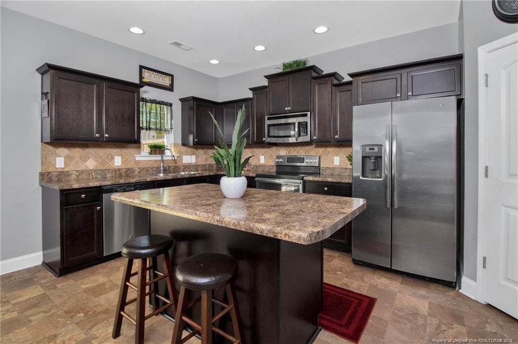 The kitchen features dark wood cabinetry complemented by a large island with seating. Stainless steel appliances are included, and a window illuminates the space. Neutral tones and a decorative plant enhance the inviting atmosphere of this functional cooking area.