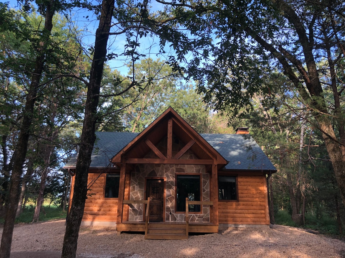 A log cabin with a stone accent facade is surrounded by tall trees, situated on a gravel path. A covered porch spans the front, featuring wooden railings. Large windows allow natural light to fill the interior, providing a cozy retreat in the woods.