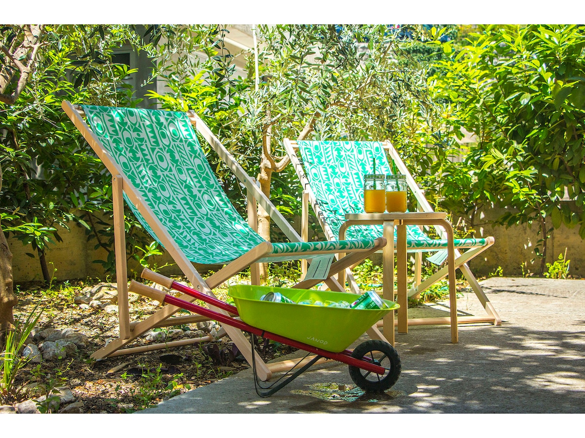 Two lounge chairs with vibrant green fabric are positioned in the garden, accompanied by a small table. A bright green wheelbarrow rests nearby, adding a touch of color to the natural setting, which features lush greenery and flowering plants.