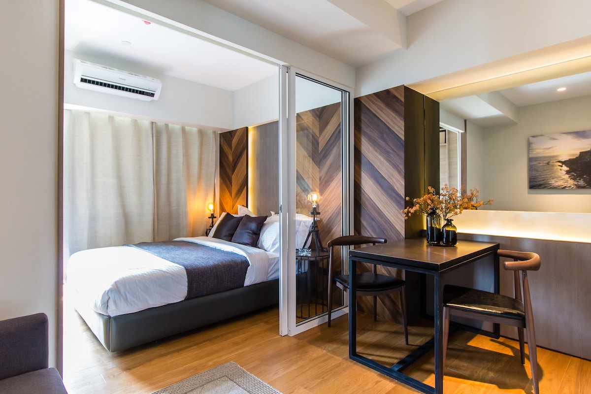 A modern bedroom area features a spacious bed with dark linens, complemented by soft lighting from lamps on either side. A small dining table with two chairs is positioned near the window, which is dressed in light curtains, allowing for a bright atmosphere.