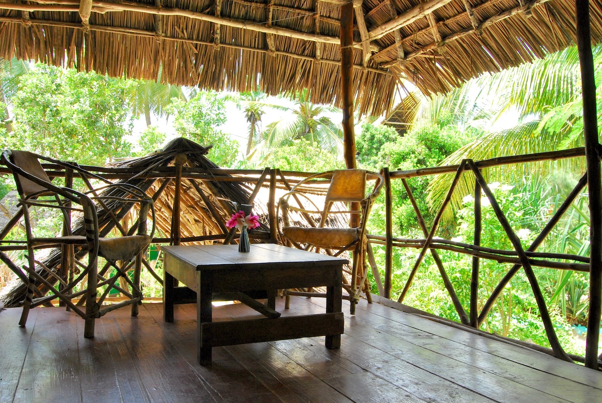 A private balcony is showcased, featuring a wooden table with a small vase of flowers. Two rustic chairs are positioned around the table, surrounded by lush greenery. The thatched roof provides a natural ambiance, enhancing the outdoor experience.