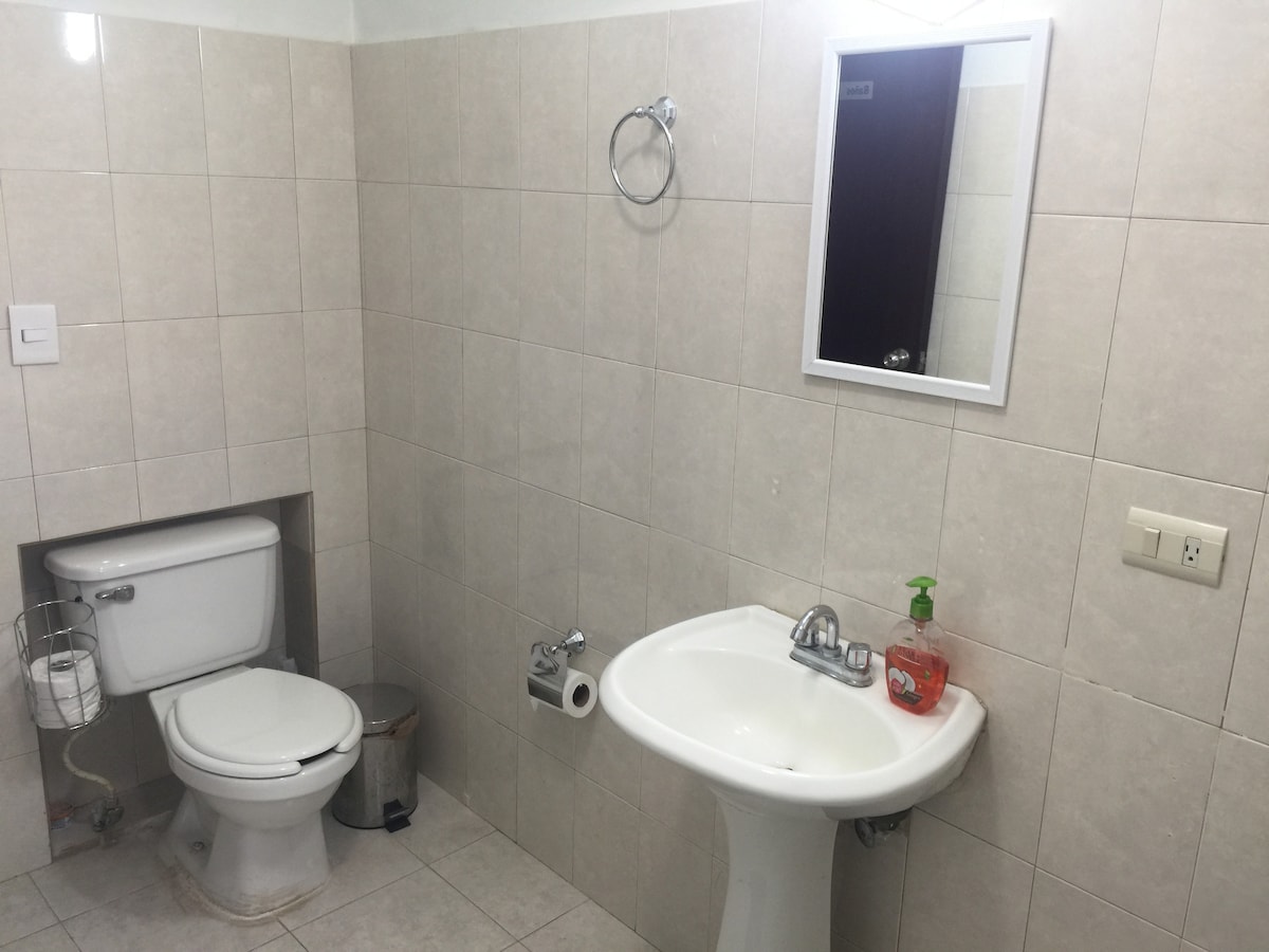 A clean bathroom is shown, featuring light-colored tiled walls and flooring. A white sink with a mirror above is positioned next to a toilet. Basic toiletries are placed on the sink, and a small wastebasket is visible in the corner.