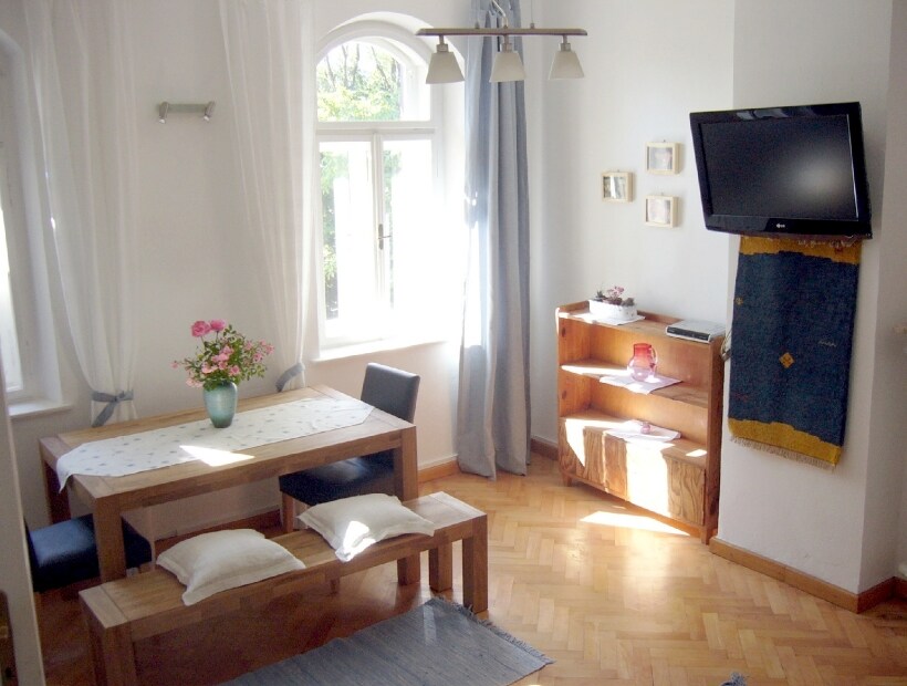 A dining area features a wooden table with simple chairs, adorned by a vase of flowers. A light-colored rug complements the warm wooden flooring. Nearby, a wall-mounted television is positioned above a wooden shelf, with natural light streaming through a window dressed in light curtains.