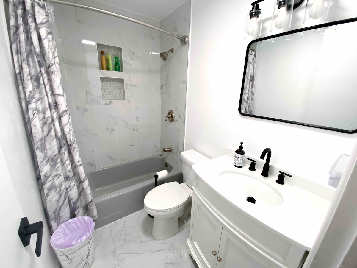A well-maintained bathroom is shown, featuring a white vanity with a round mirror and black fixtures. A bathtub with a showerhead is present, surrounded by elegant marble-like tiles. A light-colored shower curtain drapes next to the tub, and toiletries are neatly arranged on display.