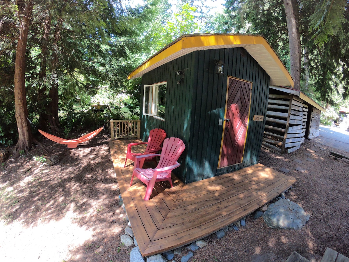 A cozy cabin is nestled among tall trees, featuring a yellow-roof and dark wooden siding. Two red chairs are positioned on the wooden deck, inviting relaxation. Nearby, a hammock is suspended between trees, enhancing the tranquil outdoor setting.