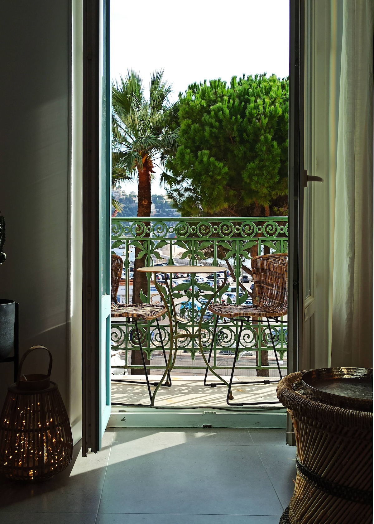 A door stands open to a balcony with a view of lush green trees and a glimpse of the sea. Two wicker chairs and a small table are situated on the balcony, inviting relaxation.