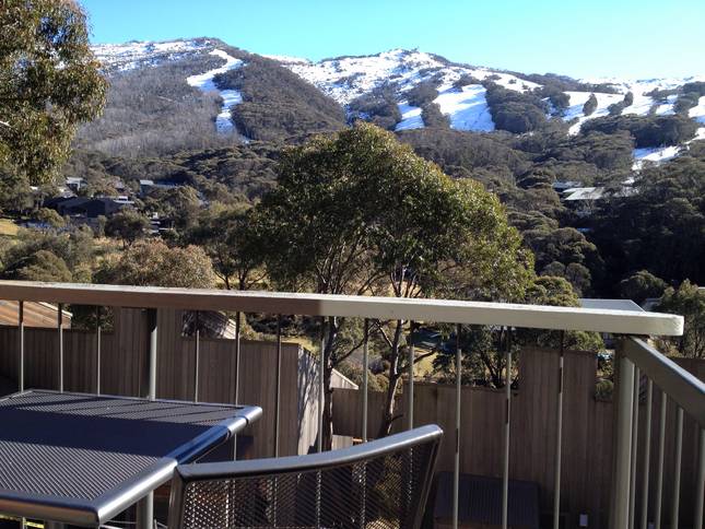 A view from a balcony captures a serene alpine landscape, showcasing snow-capped mountains and lush greenery. A sturdy railing frames the scene, while two outdoor chairs rest on the balcony, emphasizing the peaceful surroundings.