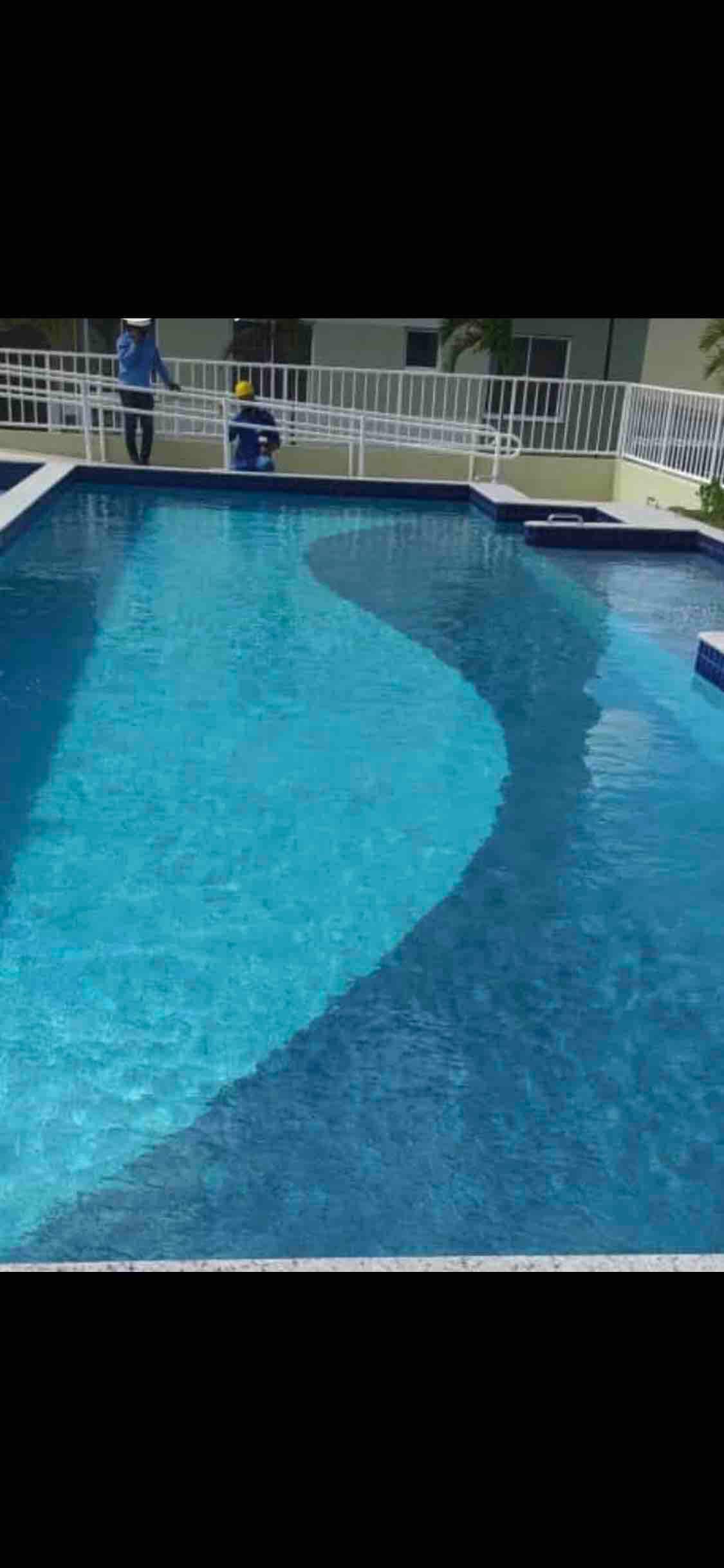 A large swimming pool features clear blue water, showcasing a gentle wave pattern on its surface. Surrounding the pool, a white fence provides an open atmosphere, while lounge chairs and poolside areas are visible for relaxation.