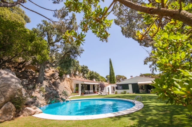A serene outdoor view features a private swimming pool surrounded by lush greenery and decorative rocks. The villa's white facade is visible to one side, while inviting shaded areas with trees create a tranquil ambiance. The vibrant blue water glistens under the clear sky.