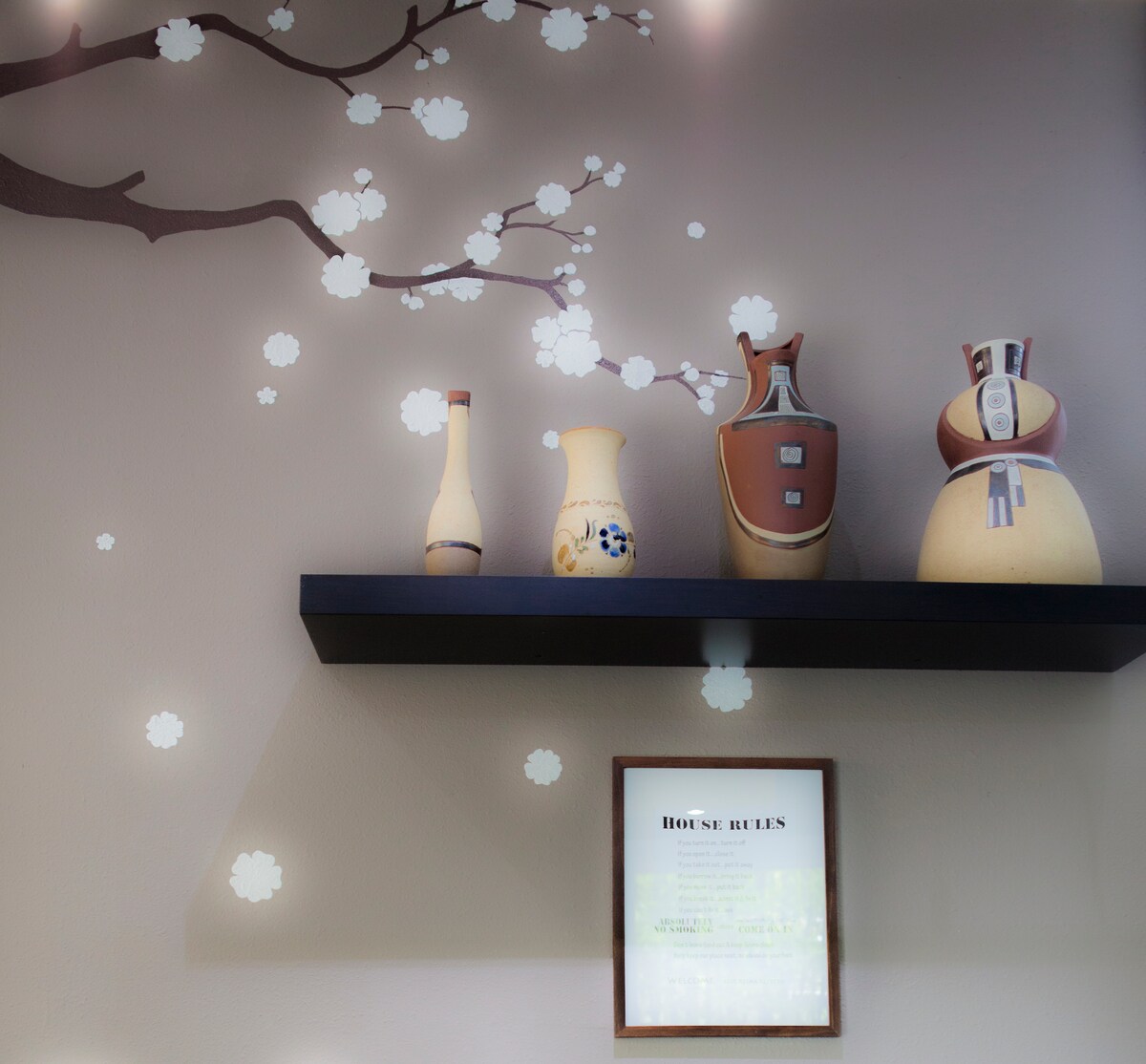 A decorative shelf is displayed against a muted wall, featuring three vases with distinct shapes and colors. Beneath them, a framed document with house rules is prominently featured. Delicate flower motifs are painted on the wall, adding a subtle nature-inspired touch.