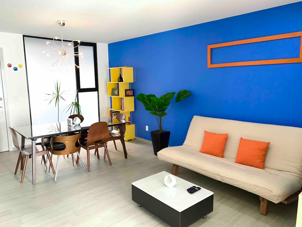 A bright living area features a blue accent wall complemented by modern furnishings. A dining table with wooden chairs is positioned beside a compact bookshelf. A light-colored sofa with orange pillows is inviting, while a potted plant adds a touch of greenery. Natural light enhances the space.