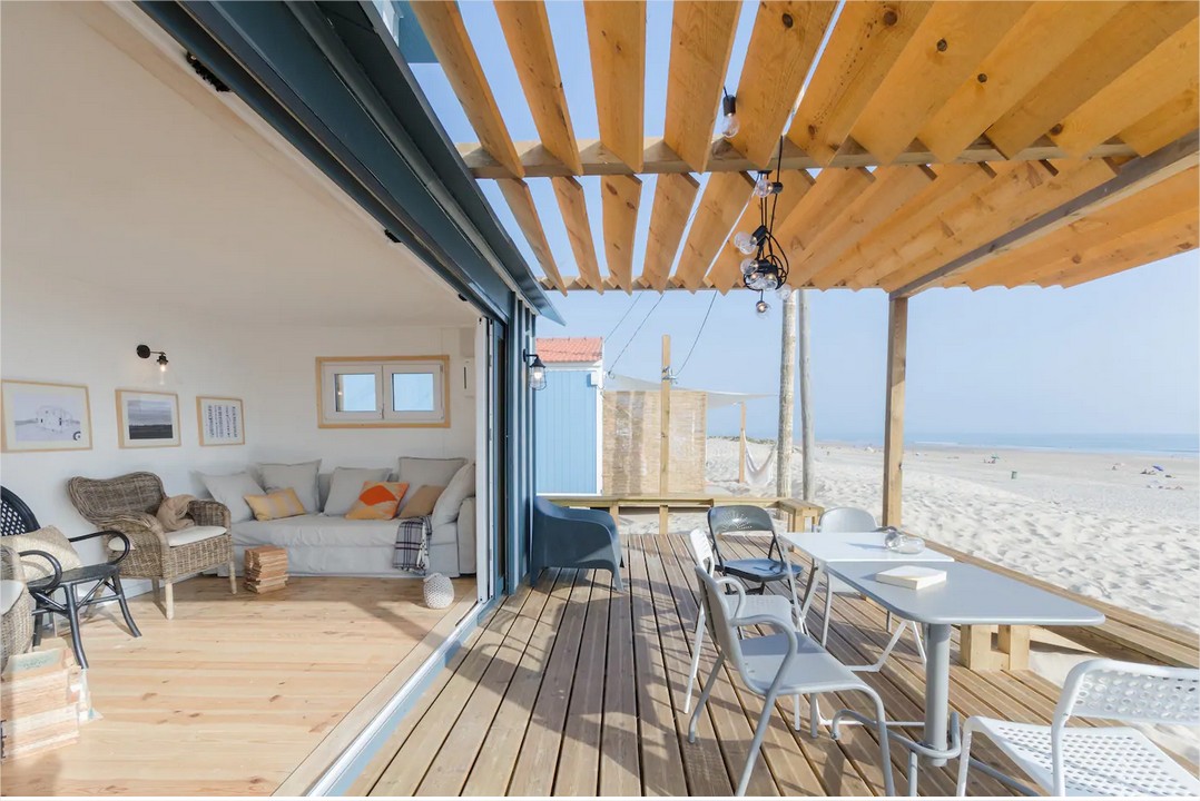 An open deck area features a wooden pergola supported by beams, accompanied by a light-filled living space. A cozy seating arrangement with cushions is complemented by a dining table set for outdoor meals. The ocean and sandy beach can be seen in the distance.