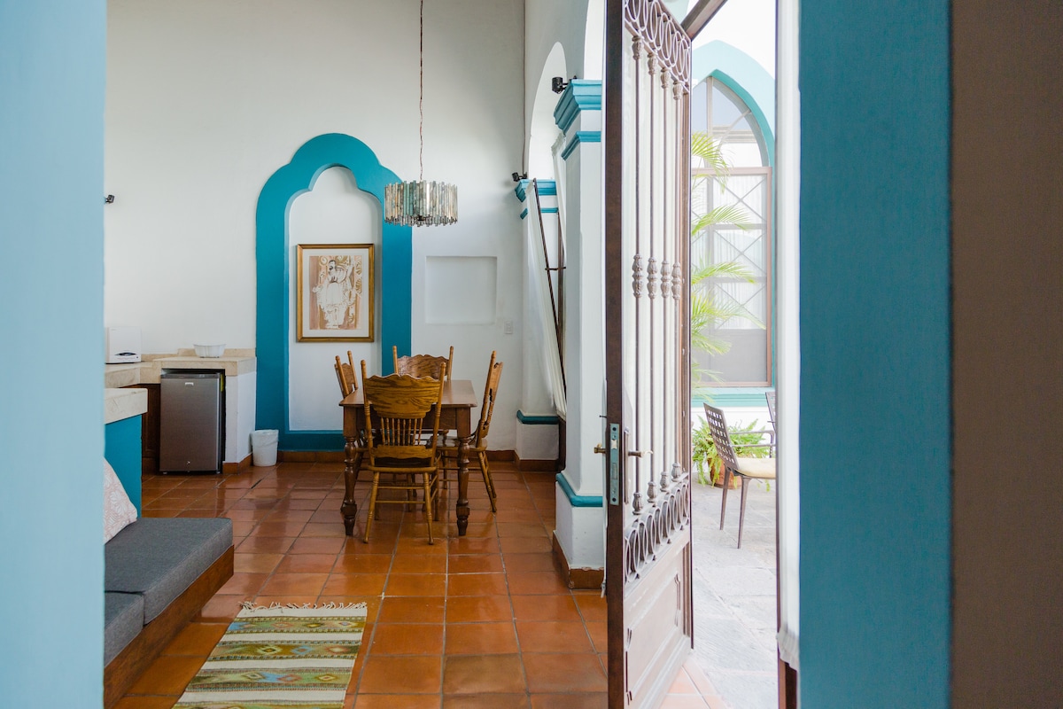 An inviting interior space features a dining area with wooden chairs around a large table. Natural light enters through the arched doorway leading to an outdoor area. A small refrigerator is visible along with a gray sofa and a decorative rug on the terracotta tile floor.