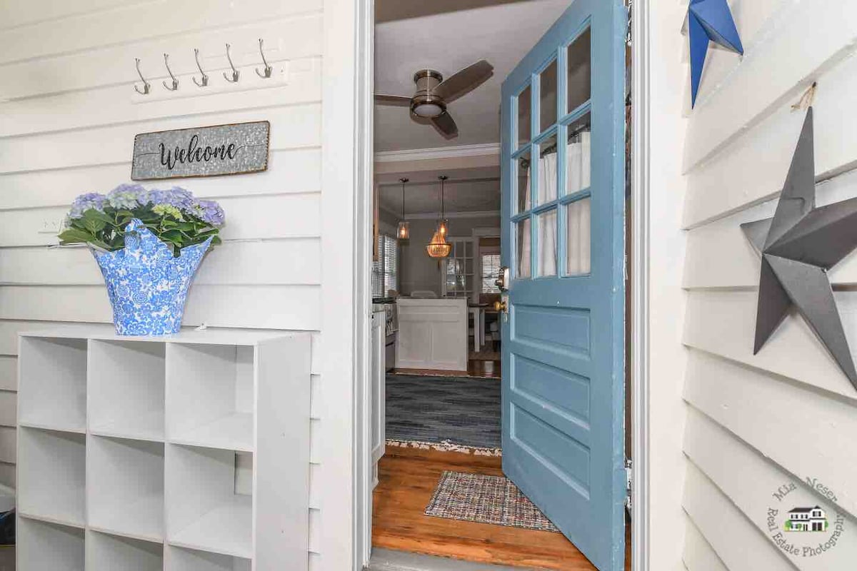 The entrance showcases a light blue door, partially open, inviting guests inside. A welcome sign and a potted plant with vibrant flowers are visible. A white storage shelf is positioned against the wall, enhancing the entryway's functionality and charm.