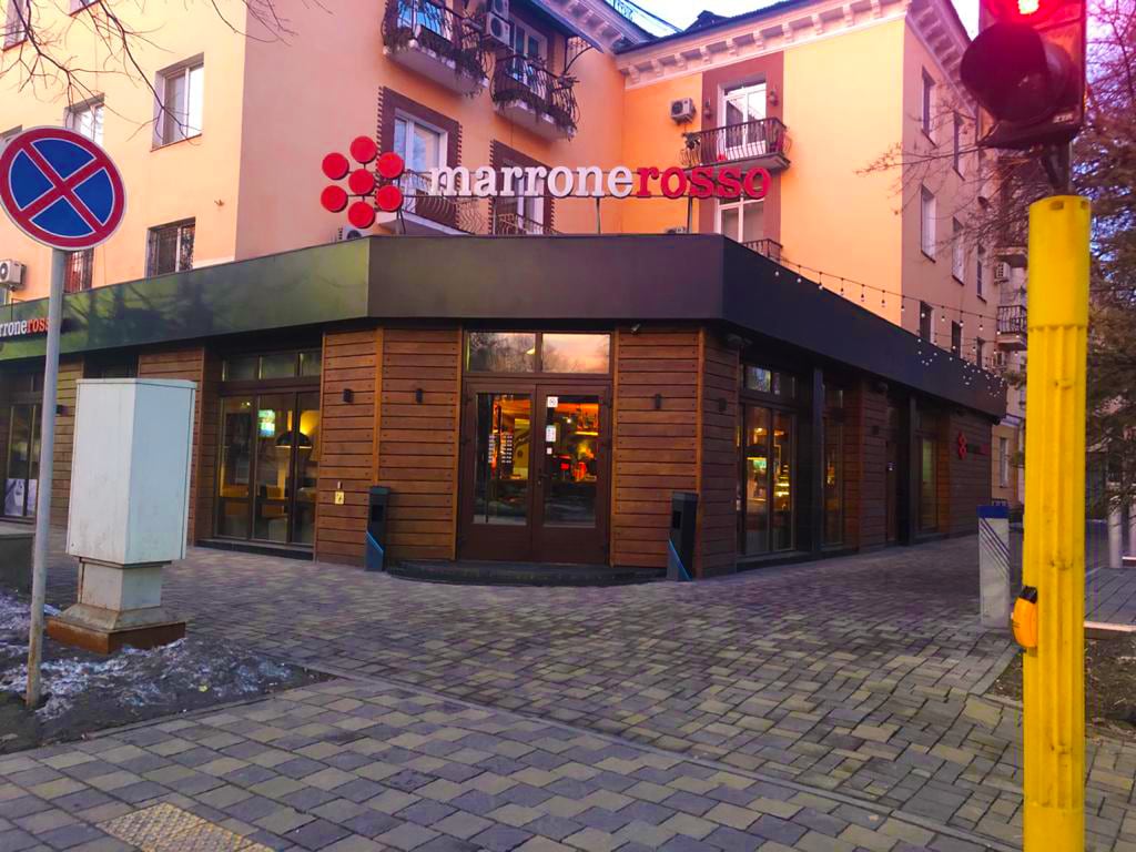 A corner building is depicted, featuring a wood-accented facade and large glass windows. The establishment, named Marrone Rosso, displays a prominent logo above the entrance. Sidewalks are paved with stones, and urban elements such as traffic signs and lampposts are visible nearby.