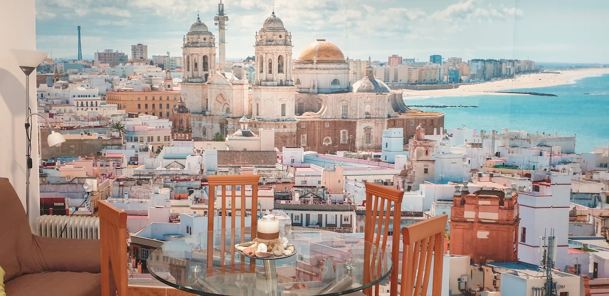 A dining area features a glass table surrounded by wooden chairs, with a picturesque wall mural displaying a panoramic view of Cádiz. The image captures the cityscape, highlighting domed buildings and the coastline against a clear sky.