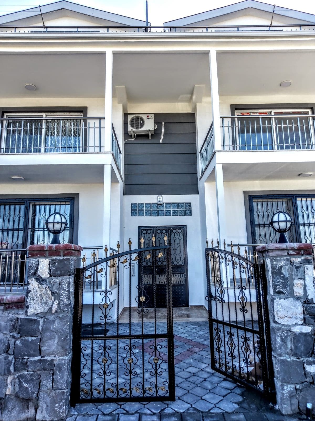 The exterior of a newly constructed building is showcased, featuring a modern design with light and dark color contrasts. A gated entrance leads to a stone pathway. Symmetrical balconies enhance the facade, while large windows allow for natural light. Air conditioning units are visible.