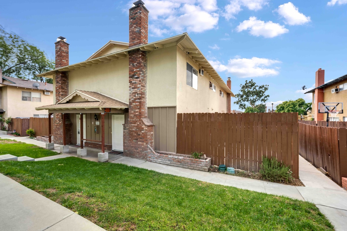 An exterior view of a two-story apartment building is presented, featuring a brick façade and a welcoming porch area. The property is bordered by a wooden fence, and the well-maintained lawn is accompanied by a pathway leading to the entrance.