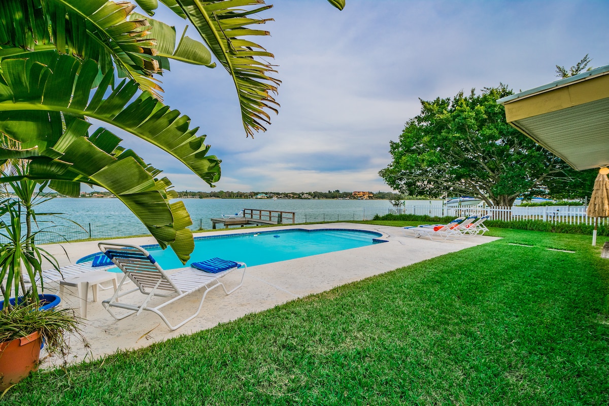 A serene outdoor area features a swimming pool surrounded by lush grass. Lounge chairs are positioned around the pool, offering views of the waterfront. Palm trees and larger greenery frame the space, creating a tranquil setting by the water.