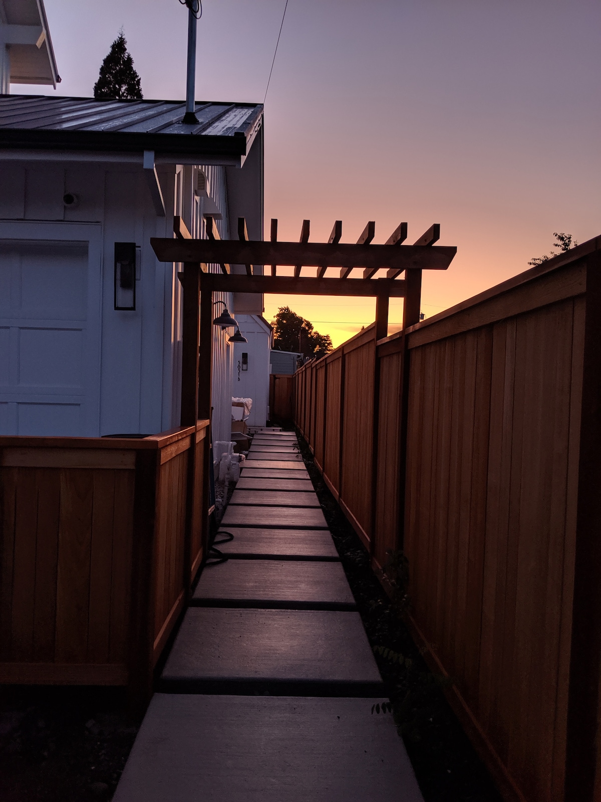 A pathway is framed by wooden fences and features a pergola overhead at sunset. The stone walkway leads to a private entrance, with soft lighting illuminating the sides, enhancing the sense of tranquility and privacy.