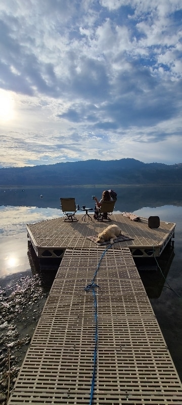 A wooden dock extends over calm water, with a person seated on a chair next to a dog resting nearby. Mountains are visible in the background under a partly cloudy sky, reflecting softly on the water's surface.