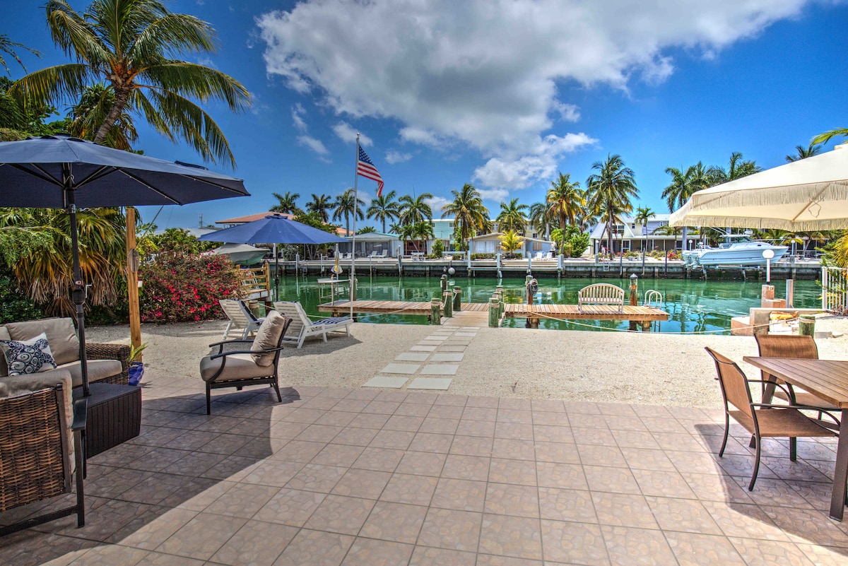 A serene outdoor space showcases a patio adorned with stylish seating and parasols, providing shade. The waterfront view features palm trees lining the canal, with boats docked in the background. Bright skies and a gentle breeze enhance the tranquil atmosphere of the area.