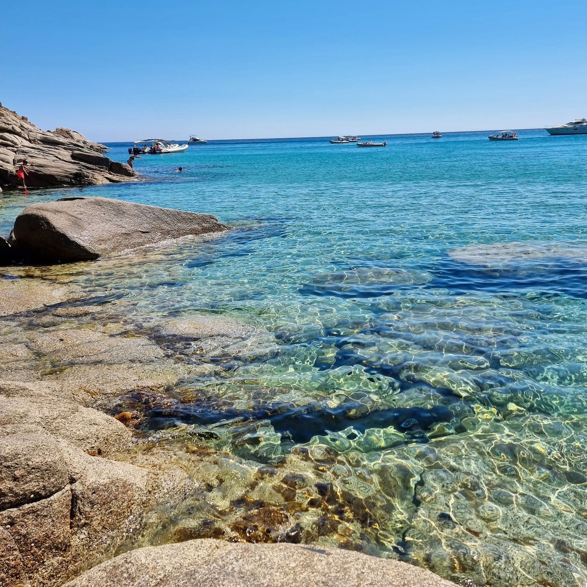 Crystal-clear turquoise waters are visible, revealing rocks beneath the surface. Small boats can be seen in the distance, with a rocky shoreline framing the scene. The sunny blue sky creates a tranquil atmosphere at this beach destination.