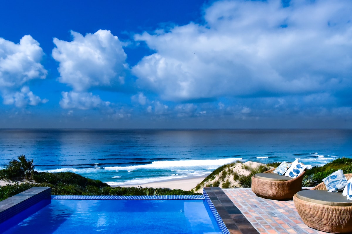 An infinity pool stretches towards the ocean, framed by lush greenery and sand dunes. Two round loungers are positioned nearby, offering a comfortable space to relax. The sky is a vibrant blue with scattered clouds, while gentle waves can be seen lapping at the shore.