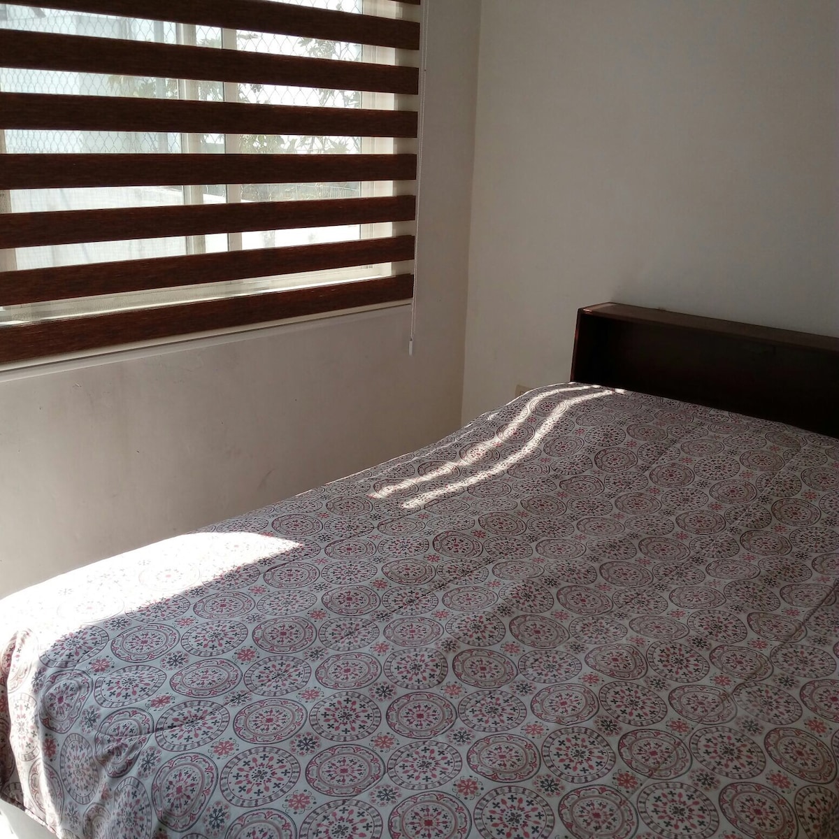 A spacious bed is shown, covered with a patterned bedspread. Natural light streams in through a window with horizontal blinds, creating a soft contrast on the wall. The simplicity of the room allows for a calm and inviting ambiance.