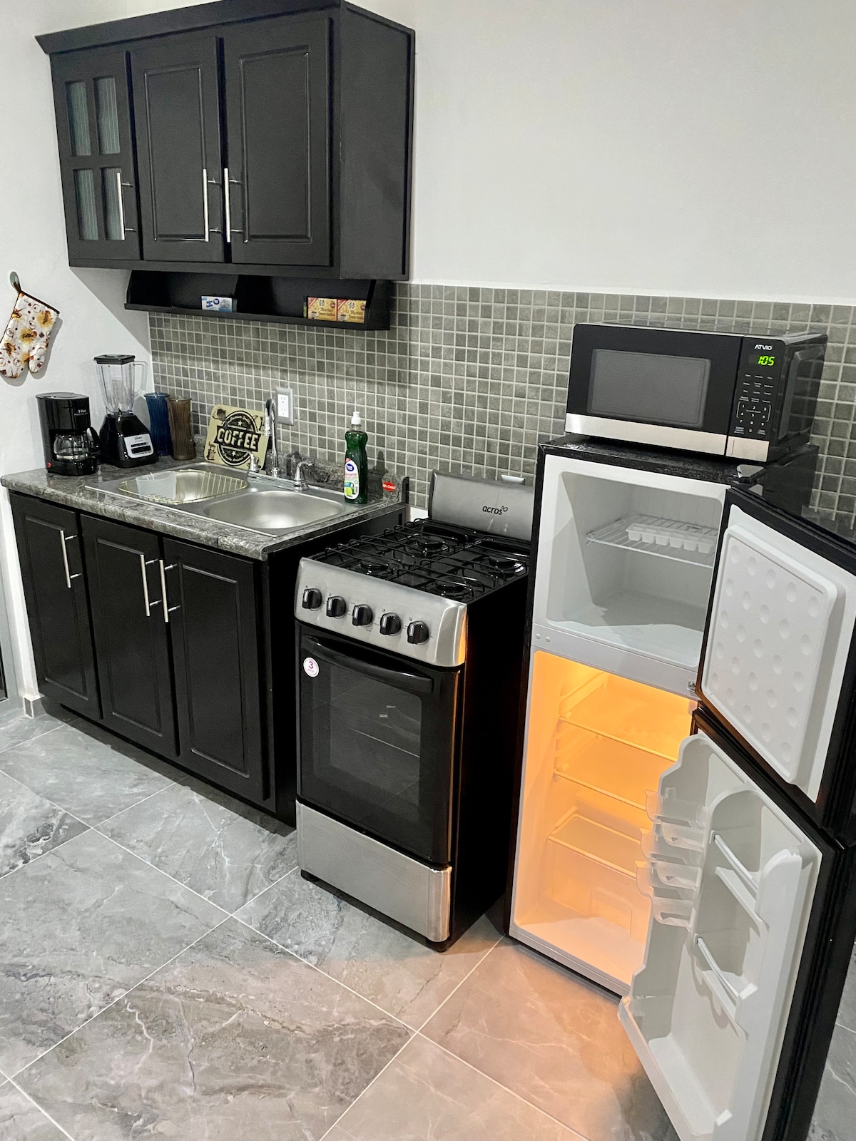 A compact kitchen space features dark cabinetry with a neutral-toned tiled backsplash. A stainless-steel stove and oven are situated next to a refrigerator with an illuminated interior. A microwave is positioned above the countertop, while a sink and small appliances are also present.