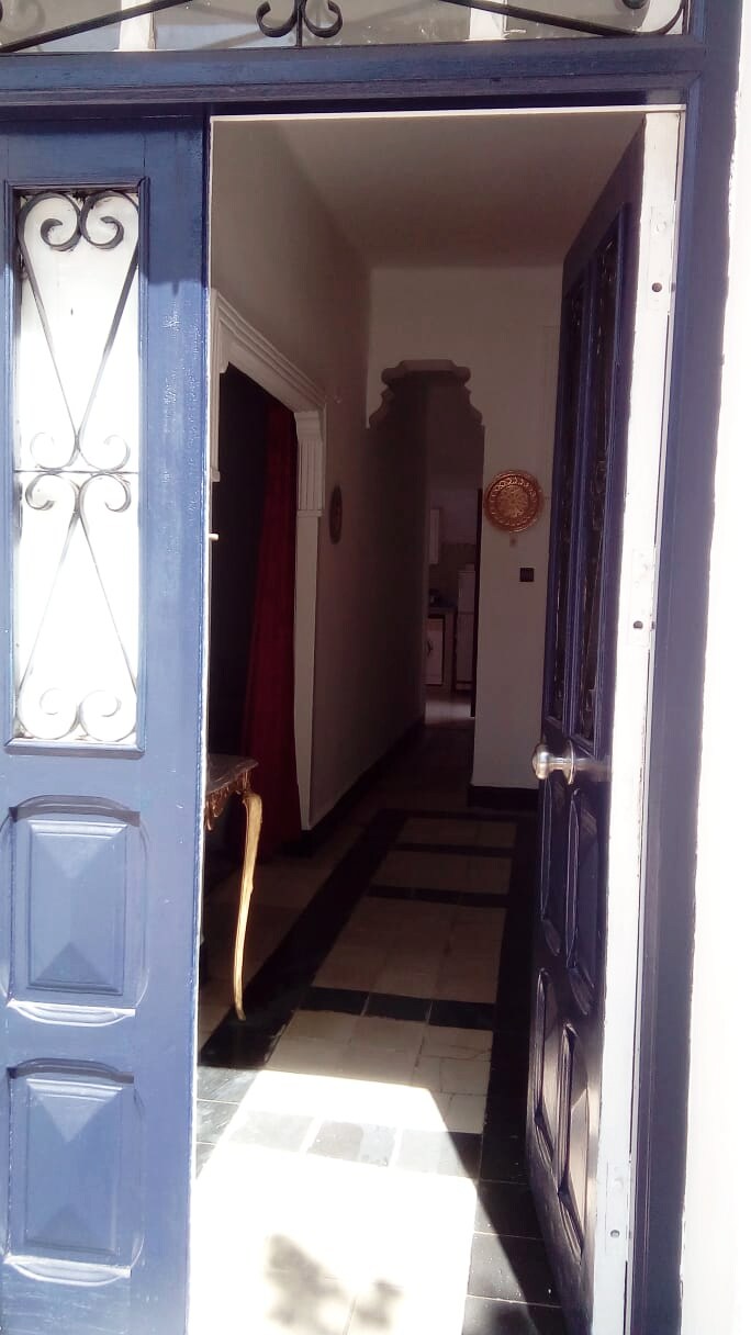 A blue front door with decorative ironwork opens to reveal a narrow hallway. The hallway features contrasting black and white tiled flooring, with warmth added by a glimpse of red drapery on one side. Light enters from the doorway, illuminating the path ahead.