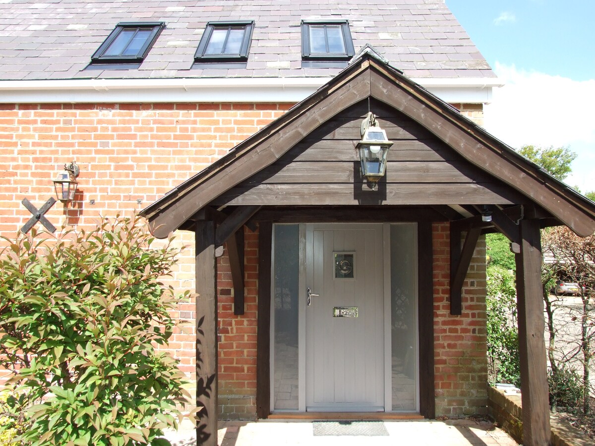 The entrance features a wooden overhang with a lantern, providing shelter. A solid grey door is framed by brick walls adorned with a lush green shrub, welcoming guests to the cottage. Above, three small windows allow natural light into the upper level.