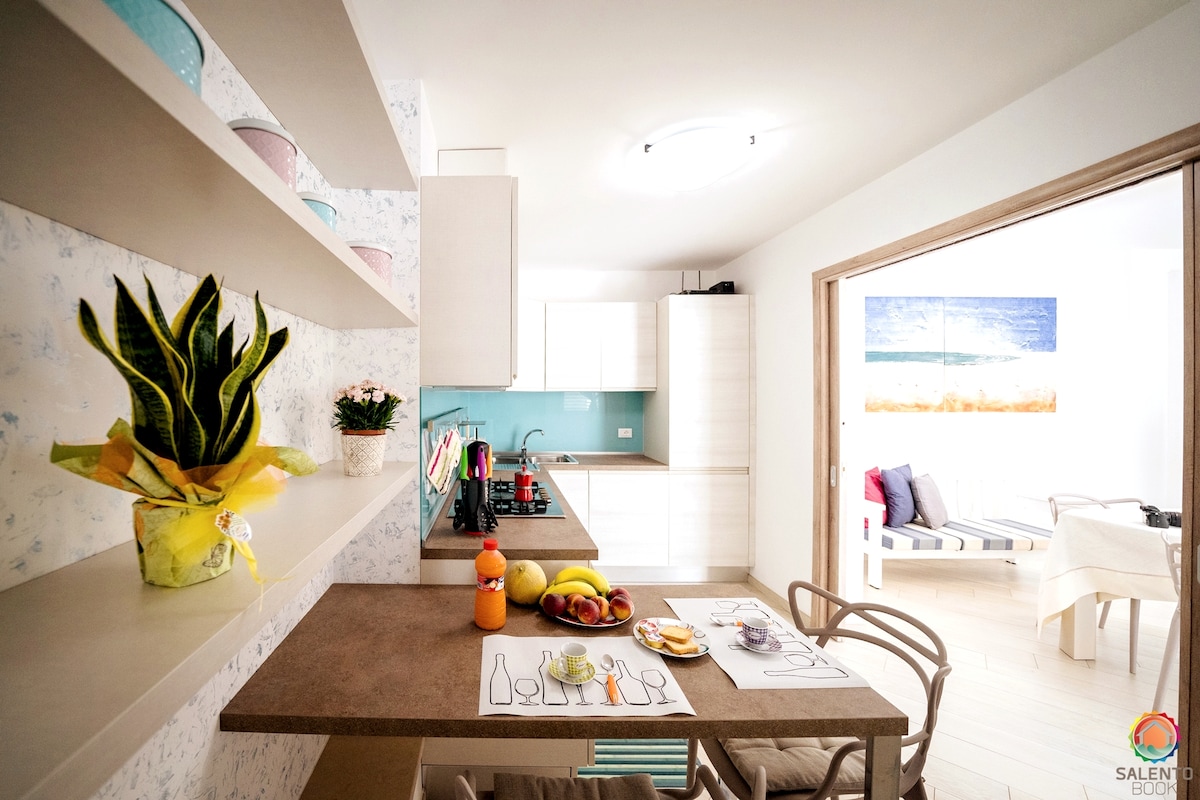 A modern kitchen area is shown, featuring sleek cabinetry and a central table set with fruits, beverages, and tableware. Decorative plants add a touch of greenery. An open doorway leads to a living space adorned with coastal-themed art.