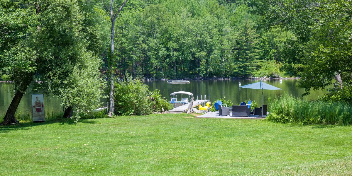 A lush green yard leads to a calm waterfront featuring a dock with seating. Bright yellow and blue chairs are positioned near an umbrella for shade. The serene surroundings are complemented by tall trees and a gentle shoreline, providing a peaceful atmosphere for relaxation.