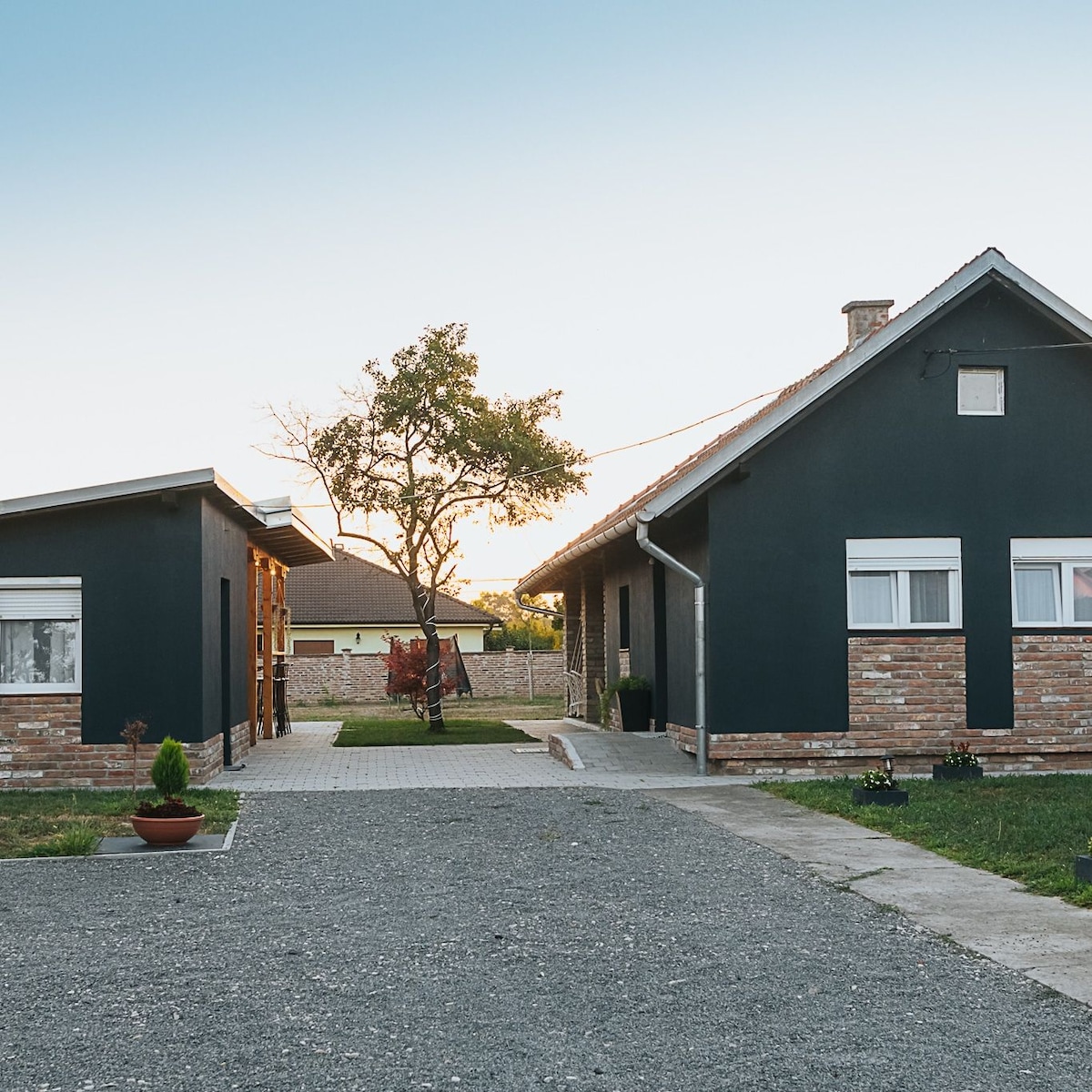 The exterior of the house is displayed, featuring a dark façade with white window frames. A gravel pathway leads through the property, flanked by greenery and a planted pot. The setting sun casts a warm glow over the area, enhancing the tranquil atmosphere.