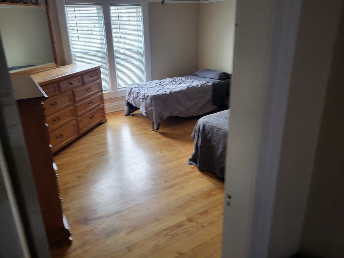 A room features two beds with neutral bedding and a wooden dresser against the wall. Natural light filters in through two windows, illuminating the warm wooden floor. The overall layout promotes a sense of openness and comfort.