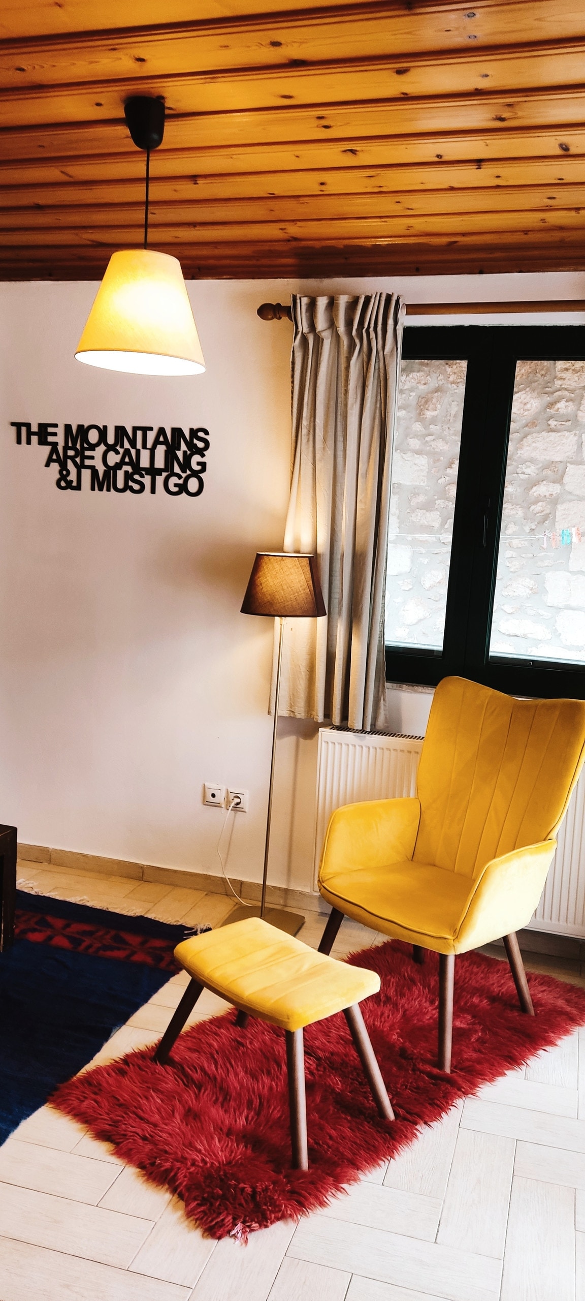 A bright yellow chair and matching footrest are positioned atop a plush red rug. A floor lamp and a pendant light illuminate the space. Wall decor features text centered on a white wall, and a large window with light curtains allows natural light to fill the room.