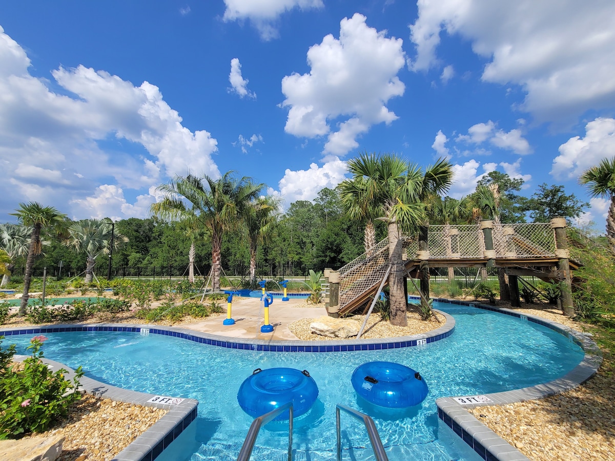 A winding lazy river surrounded by lush greenery features several palm trees and a wooden climbing structure. Two bright blue inner tubes float invitingly on the clear water, and a decorative pathway leads along the water's edge.