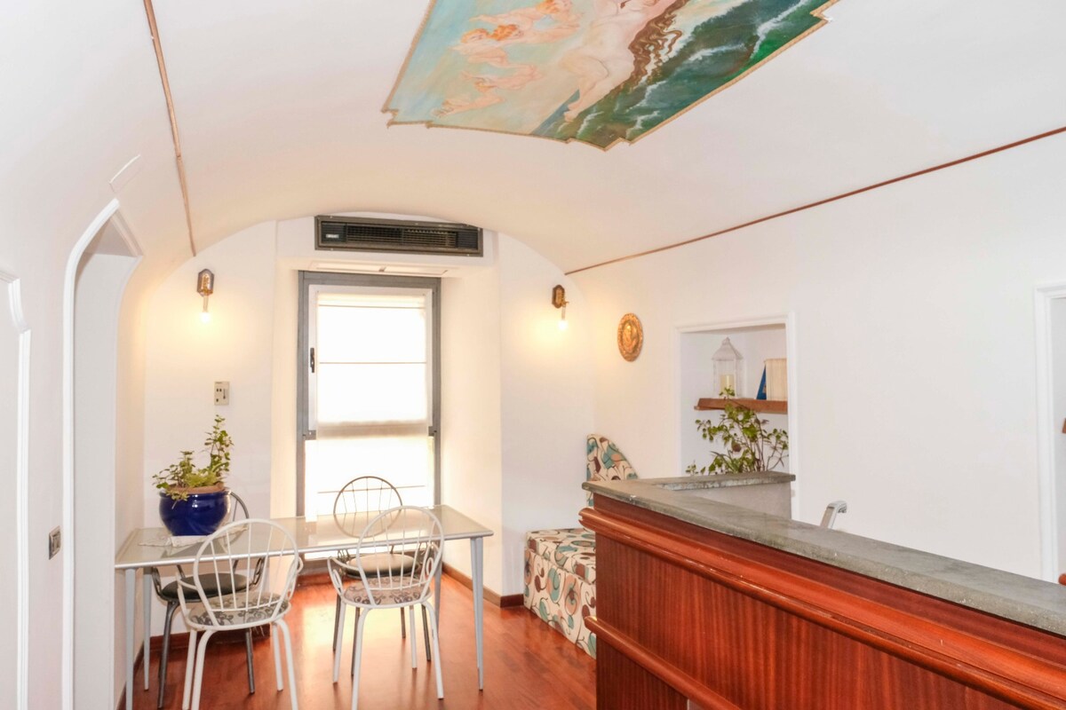 A cozy dining area is set with a simple table and four metal chairs, complemented by a colorful couch adorned with floral patterns. A decorative mural graces the arched ceiling, while natural light filters through the window, enhancing the inviting space.