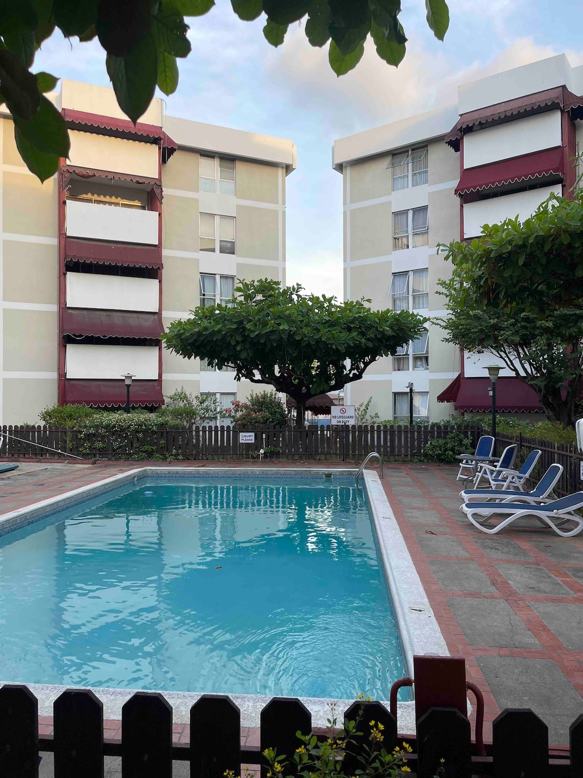 A swimming pool is centrally located between two apartment buildings, surrounded by greenery. Comfortable lounge chairs are placed nearby, and a large tree provides shade. The area features a fence and well-maintained landscaping, creating a relaxing atmosphere.