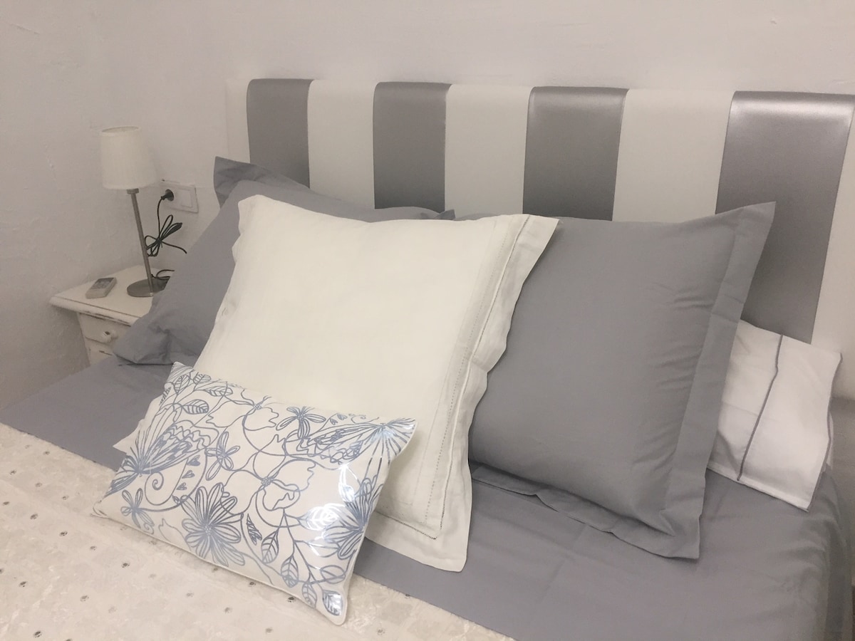 A spacious bed is presented with neatly arranged decorative pillows in shades of gray and white. A bedside lamp provides subtle illumination, and a nightstand houses a simple alarm clock. The bed linens exhibit a soft texture against a light-colored wall.