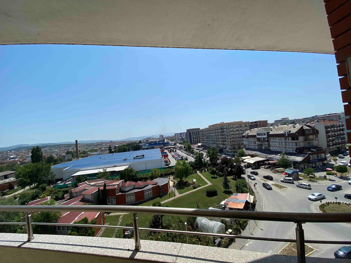 A panoramic view is captured from a seventh-floor balcony. Urban landscapes, including buildings, roads, and greenery, are visible under a clear blue sky. The railing emphasizes the elevated perspective, enhancing the openness of the scene.