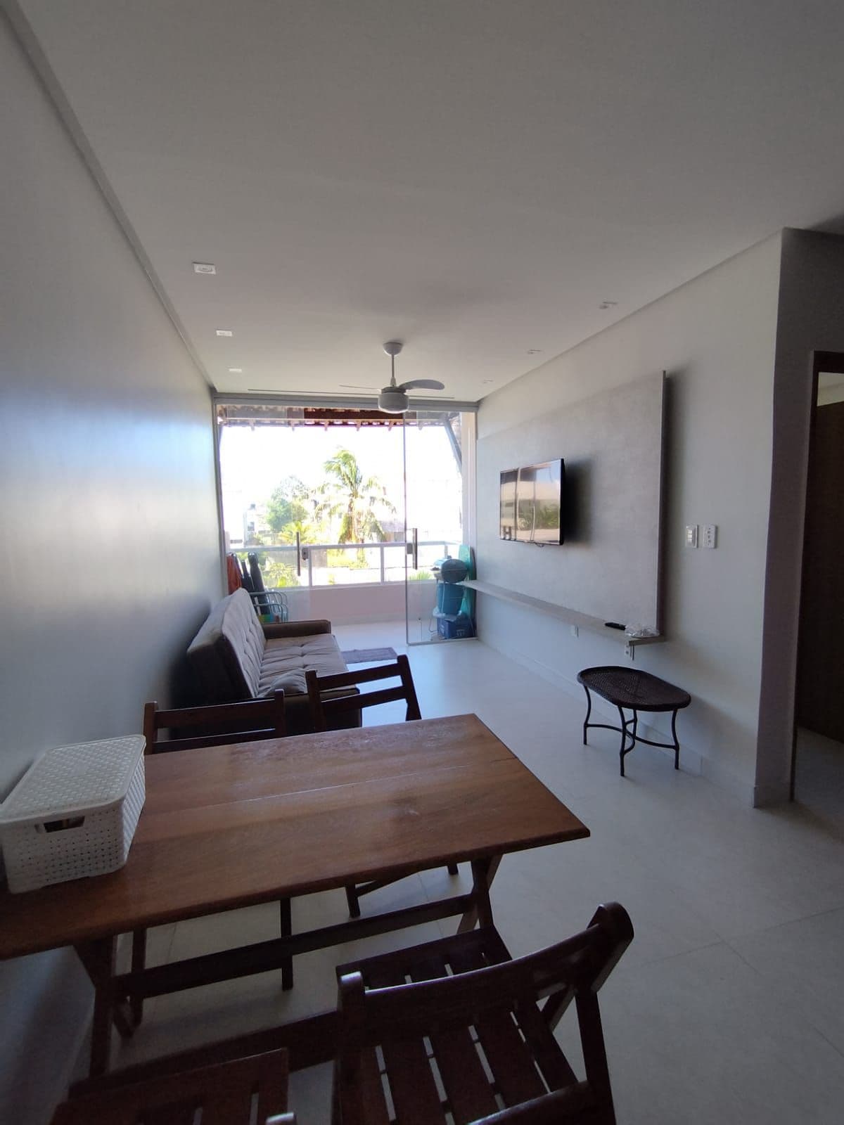 A modern living area is presented with a wooden dining table and chairs. A comfortable sofa is placed against one wall, while a mounted television is installed opposite. Natural light illuminates the space through a large window, offering views of the surrounding greenery.