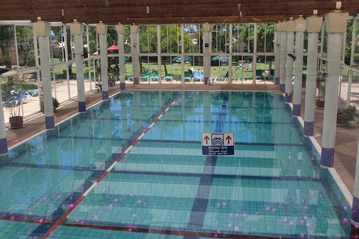 An indoor heated swimming pool is framed by tall glass windows, providing views of the surrounding garden. The pool features a clear blue tile design, marked lanes, and safety signage. Lounge chairs are visible in the background, arranged around the pool area.