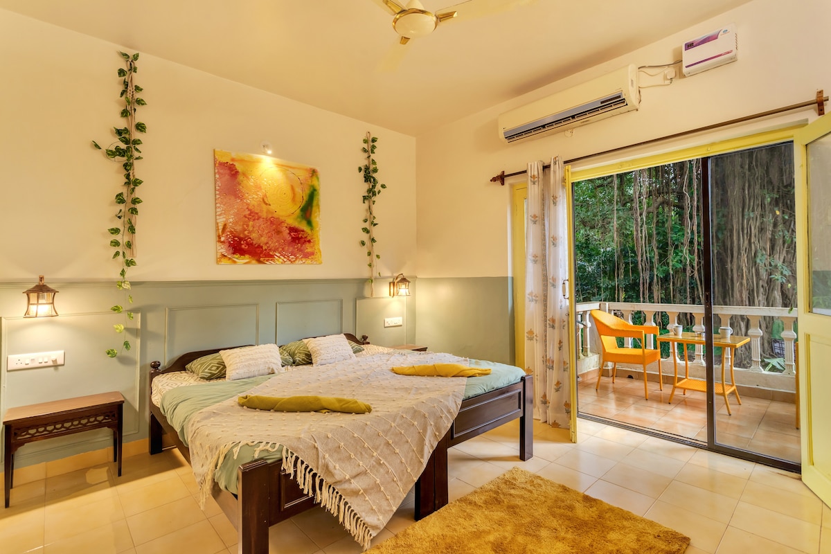 A spacious bedroom features a king-size bed adorned with a decorative throw, complemented by soft pillows. Large glass doors open to a balcony with outdoor seating, surrounded by greenery. The room is softly lit, highlighting decorative wall plants and vibrant artwork.