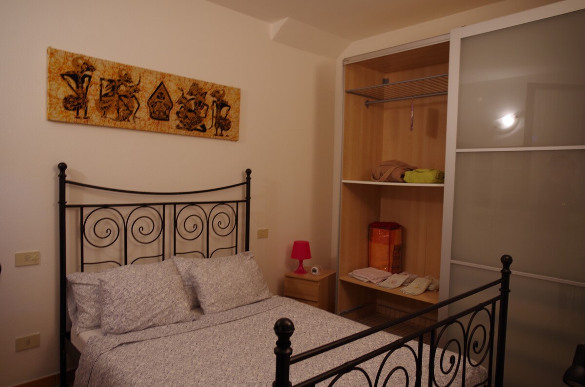 A cozy bedroom features a metal frame bed with soft bedding and decorative wall art above. A bedside table with a small lamp is positioned next to the bed. A sliding door wardrobe is visible, showing shelves and folded linens inside.