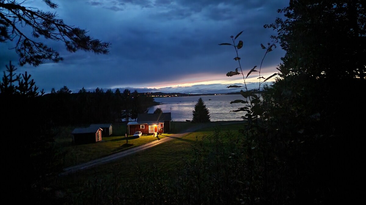 A charming house is seen against a twilight sky, with warm light glowing from its windows. A pathway leads from the house toward the water, framed by trees and grass, while the distant shoreline is illuminated by soft reflections from the water.
