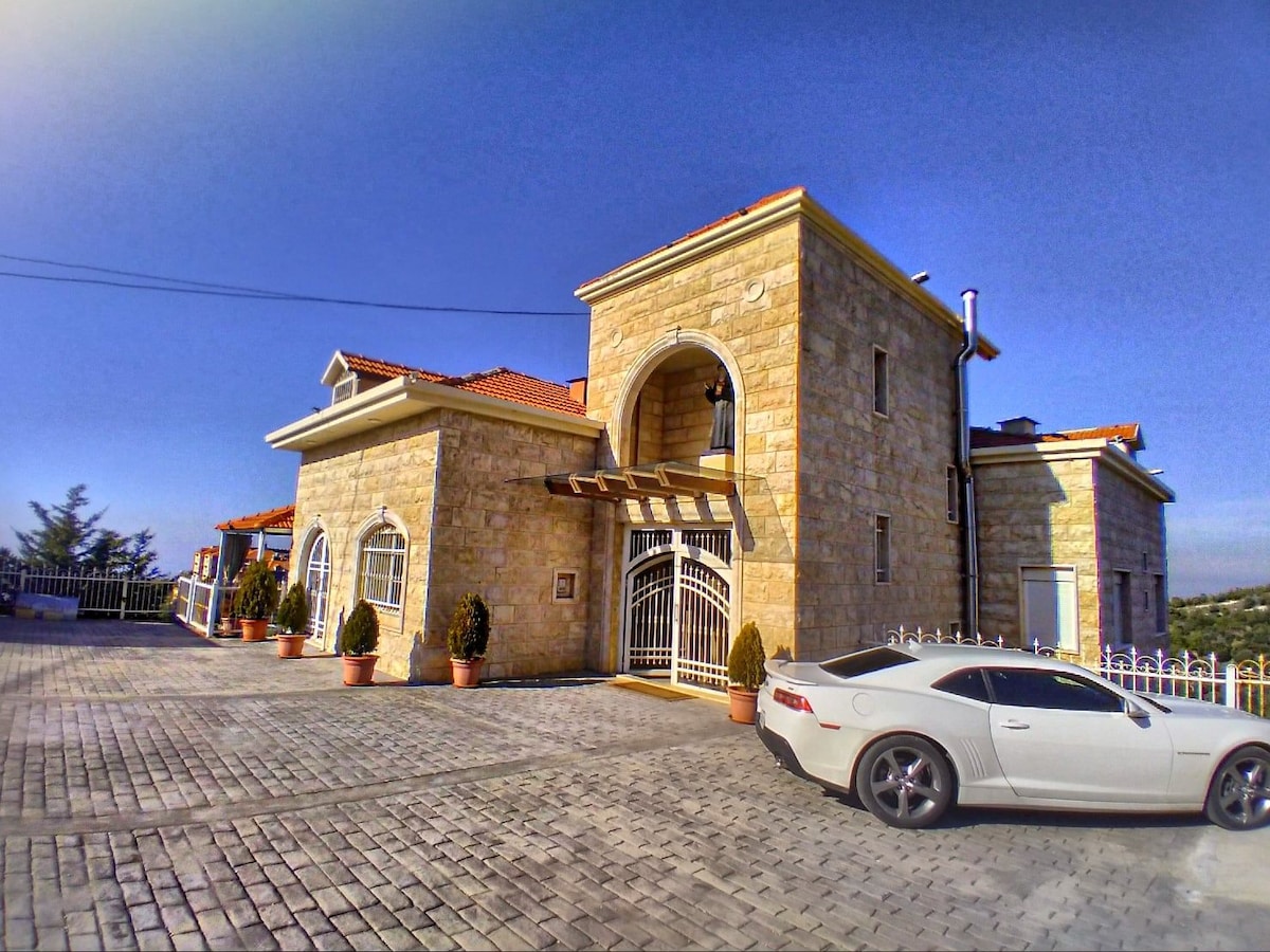 A charming stone house features a welcoming entrance with an arched doorway and decorative windows. Potted plants line the front, leading to a paved driveway where a vehicle is parked. The structure is set against a clear blue sky, highlighting its spacious and inviting design.