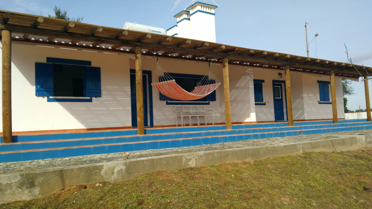 A single-story house features a covered porch with wooden beams. A colorful hammock is suspended between two posts. The façade includes blue window shutters and a blue trim along the bottom. A small white table and chairs are placed under the shelter.