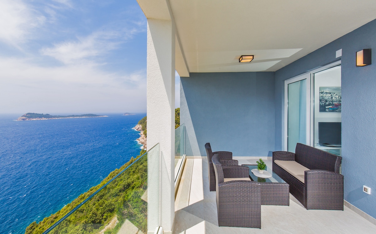 A spacious terrace features a seating area with modern, dark wicker furniture and a small table. Clear glass railings provide unobstructed views of the expansive sea and coastline, inviting relaxation in a serene outdoor environment.